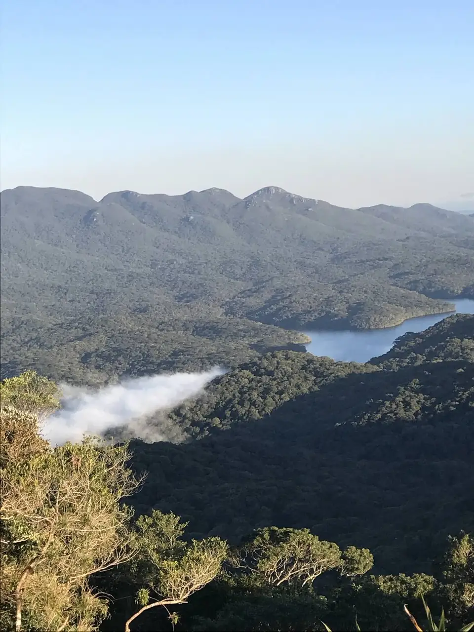 Represa entre montanhas cobertas de mata, com um fiapo de nuvem no vale